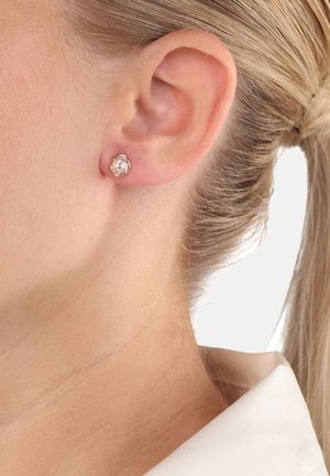 Primer plano de la oreja de una mujer rubia que lleva un pequeño pendiente de plata con una gema clara en el centro, con el cabello recogido y un cuello blanco visible.