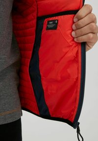 Red insulated jacket interior with quilted texture. One black inner pocket with label visible. Zippered closure and navy lining detail.