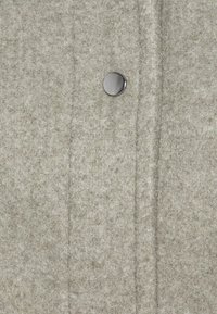 Harmaa villatakki, jossa on hienovarainen kuvio ja etuosassa metallinen neppari sekä pystysuorat saumalinjat.