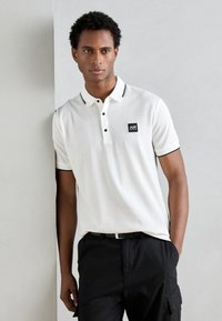 Polo bianco con colletto nero, apertura a tre bottoni e patch con logo. Abbinato a pantaloni cargo neri con tasche laterali.