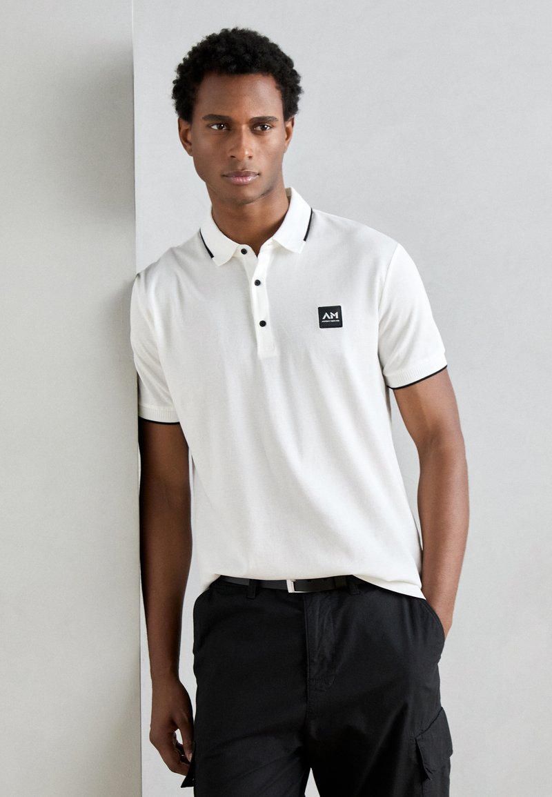 Polo bianco con colletto nero, apertura a tre bottoni e patch con logo. Abbinato a pantaloni cargo neri con tasche laterali.