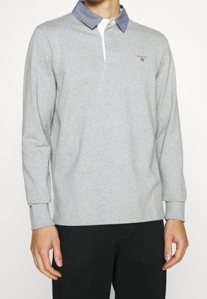 Homme portant une chemise à manches longues gris clair avec un col bleu par-dessus un sous-vêtement blanc et un pantalon noir, debout devant un fond blanc.