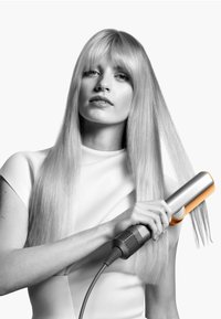 Haarglätter mit silbernem Gehäuse und orangefarbenen Akzenten, gehalten von einem Modell mit langen, glatten, hellblonden Haaren. Strukturiertem Griff und sichtbarem Stromkabel.