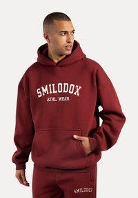 Sudadera burdeos con bolsillo delantero y puños acanalados. Presenta el texto blanco "SMILODOX ATHL. WEAR" en el pecho. Material suave de mezcla de algodón.