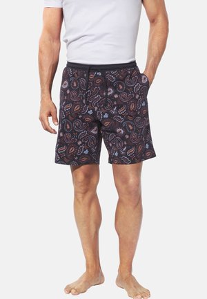 Man wearing black shorts with multicolored paisley pattern and drawstring waist, paired with a white short-sleeve shirt, barefoot.
