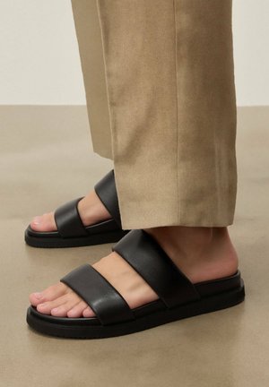 Füße in schwarzen Leder-Sandalen mit zwei Riemen, kombiniert mit beigefarbenen Hosen mit weitem Bein, stehen auf einem glatten Boden.