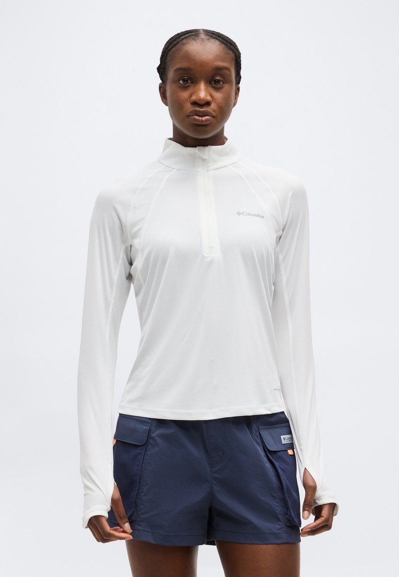 Γυναίκα φορώντας λευκό μακρυμάνικο ζιπ-απ τοπ Columbia και σκούρο μπλε cargo σορτς με τσεπάκια από δίχτυ, στέκεται μπροστά σε λευκό φόντο.