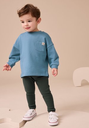 Dijete u plavoj majici dugih rukava, tamnozelenim hlačama i bijelim tenisicama stoji na bež geometrijskim oblicima.