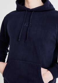 Hanorac din fleece bleumarin cu șnur, buzunar tip cangur și logo embossat pe piept. Prezintă o textură moale și manșete ajustate.