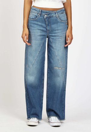 Frau trägt blau-weitgeschnittene Jeans mit mittlerer Leibhöhe, diagonalen Nahtdetails, leichtem Used-Look, rohem Saum und weißen Turnschuhen.