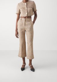 Kvinna som bär en beige och flerfärgad tweed kort jacka och matchande högmidjade, korta flarebyxor tillsammans med svarta spetsiga klackar.