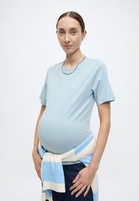 Ανοιχτό μπλε κοντομάνικο μπλουζάκι με εφαρμοστή γραμμή, φορεμένο πάνω από ένα πουλόβερ με ριγέ σχέδιο σε ανοιχτό μπλε και κρεμ αποχρώσεις, δεμένο στη μέση. Σκούρο μπλε τζιν παντελόνι.