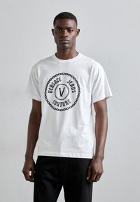 T-shirt en coton blanc avec un motif central noir portant l'inscription "Versace Jeans Couture" entourée d'un motif de chaîne. Manches courtes, col rond.