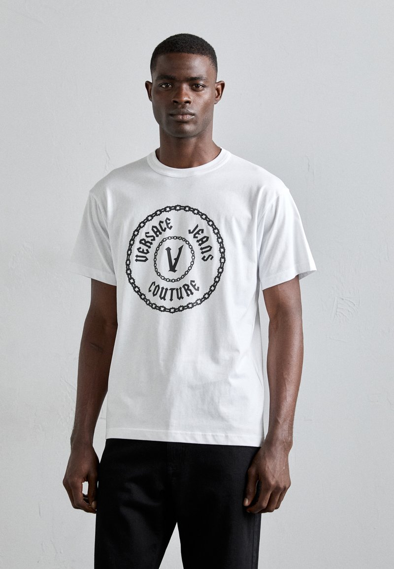 T-shirt en coton blanc avec un motif central noir portant l'inscription "Versace Jeans Couture" entourée d'un motif de chaîne. Manches courtes, col rond.