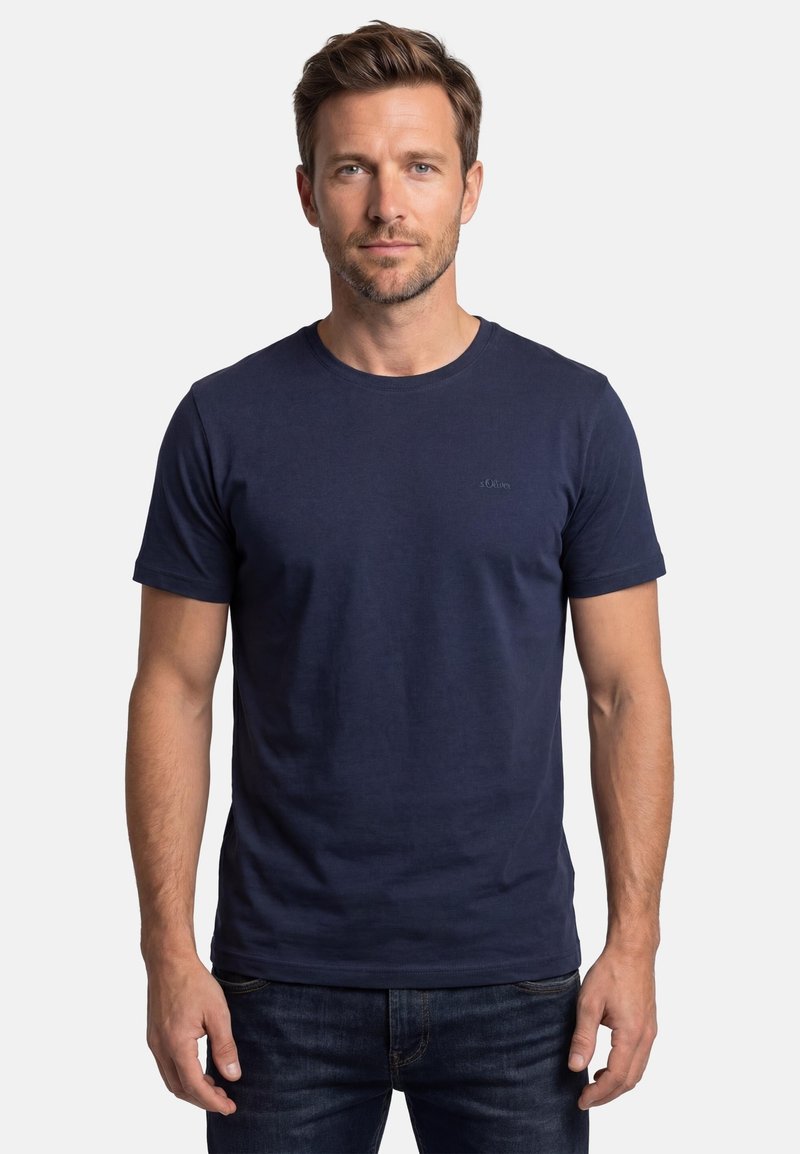 Homme aux cheveux bruns courts et à la barbe portant un t-shirt bleu marine uni et un jean foncé, debout devant un fond blanc.
