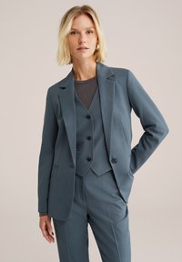 Tyrkysový oblek na míru s jednoradým sakem, odpovídajícím vestou se třemi knoflíky a úzkými kalhotami. Hladká tkanina a strukturovaný design.