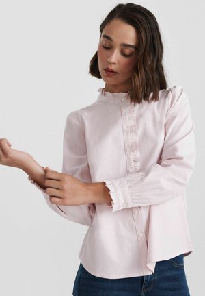 Femme ajustant le poignet d'un chemisier rose clair à volants avec des boutons, portant un jean bleu foncé, sur un fond clair uni.