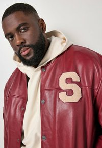 Baardige man met een crèmekleurige hoodie onder een rode leren jas met een grote beige "S"-applicatie op de borst, tegen een effen lichte achtergrond.