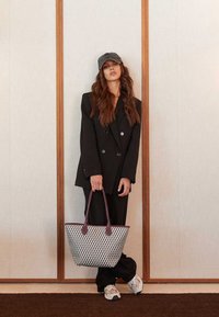 Zwarte double-breasted blazer over bijpassende broek, met een geweven wit en zwarte tote bag met bordeauxrode handvatten, en een grijze pet.