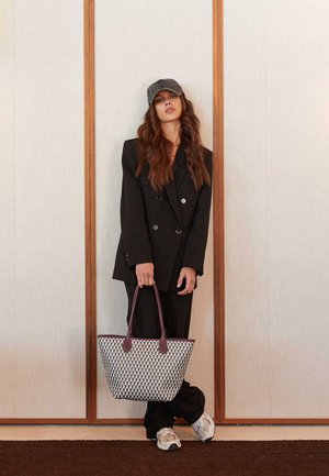 Crni dvoredni sako preko odgovarajućih hlača, drži pletenu bijelu i crnu torbu s bordo ručkama i sivu kapu.