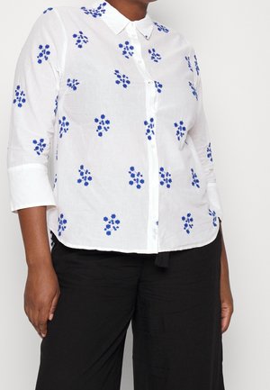 Camisa blanca de botón con bordado floral azul, mangas largas con puños doblados y un cuello clásico. Hecha de una tela ligera.
