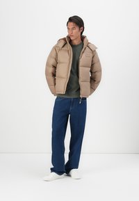 Puffervest i lys brun, med hætte, frontlynlås og boxy form. Kombineret med mørkeblå jeans og hvide sneakers.
