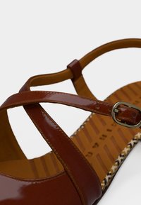 Καφέ δερμάτινο σανδάλι με διασταυρούμενα λουριά, μεταλλική αγκράφα και υφασμένο πέλμα με διακοσμητική άκρη.