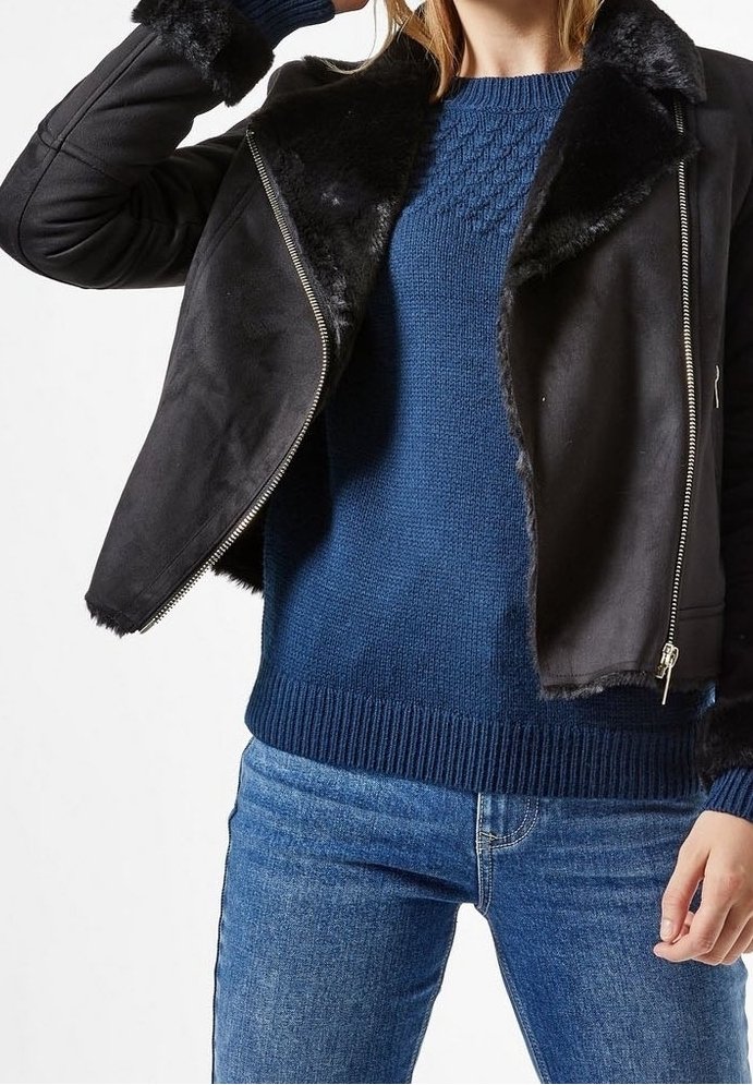Giubbotto in finta pelle nera con colletto peloso, chiusura con zip, abbinato a un maglione di maglia blu testurizzata e jeans blu.