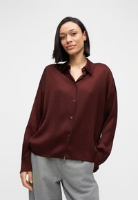 Chemise boutonnée bordeaux avec col, coupe ample et manches longues, associée à un pantalon gris ; texture lisse et détails discrets aux boutons.