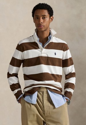 Joven con camisa de rugby de manga larga a rayas marrones y blancas sobre camisa celeste con botones, pantalones beige, de pie con las manos en los bolsillos.