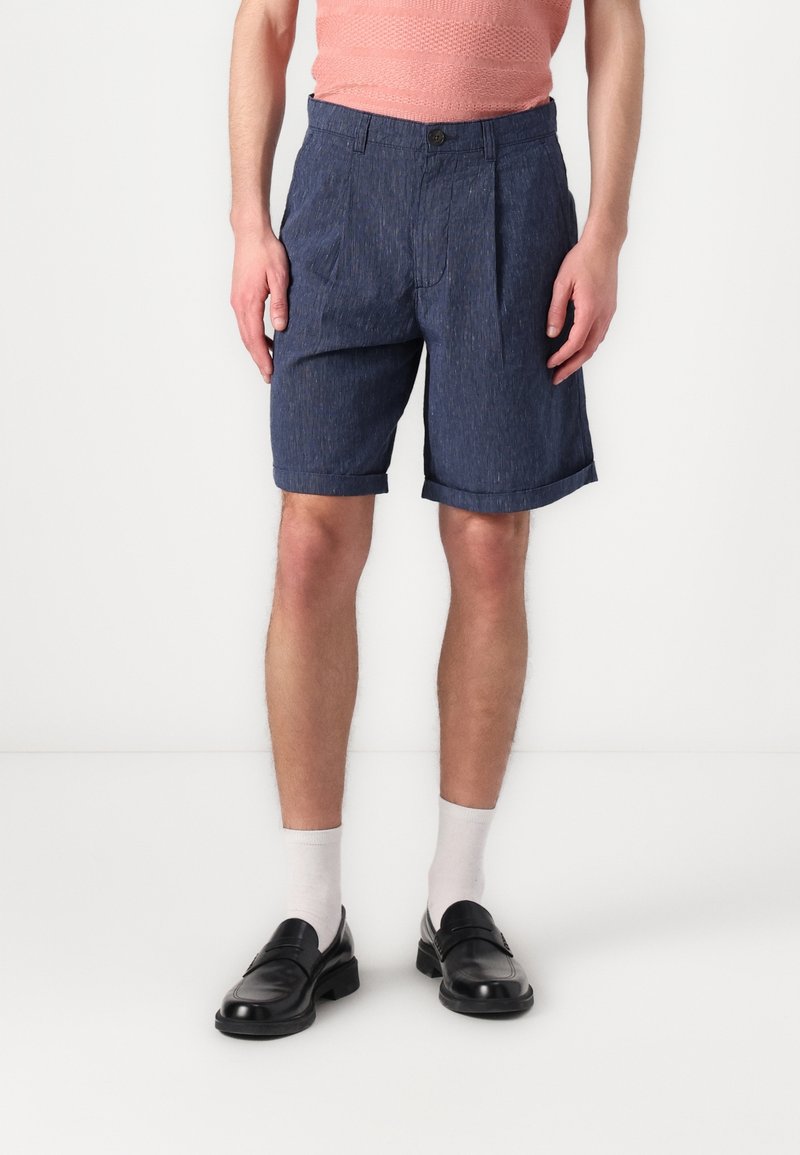 Homme portant un short bleu marine avec ourlet retroussé, des chaussettes blanches, des mocassins noirs et un haut texturé corail, sur fond blanc.