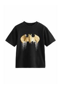 Camiseta de algodón negra con un gran logo de Batman en lentejuelas doradas. El logo presenta detalles goteantes, tiene mangas cortas y un diseño de cuello redondo.