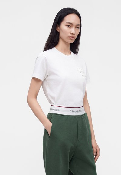 Femme aux longs cheveux noirs portant un t-shirt blanc et un pantalon vert avec une ceinture blanche portant la mention DSQUARED2, debout avec une main dans la poche.