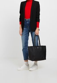 Sac fourre-tout noir en matériau synthétique avec des poignées doubles, texture lisse et petit logo. Porté avec un haut rouge, un blazer noir et un jean bleu.