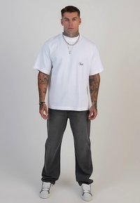 Baltas medvilninis trumpomis rankovėmis marškinėlis su mažu logotipu kairėje pusėje, derinamas su tamsiai pilkais džinsais ir baltais sportbačiais.