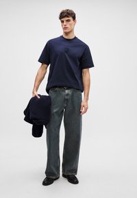 Ναυτικό μπλε βαμβακερό t-shirt με στρογγυλή λαιμόκοψη και λογότυπο μπροστά, συνδυασμένο με φαρδιά τζιν ελαφρού ντενίμ και μαύρα παπούτσια, κρατώντας ένα ναυτικό φούτερ.