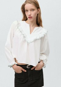 White blouse with a V-neck, frosted green ruffle trim, and gathered sleeves. Paired with black denim shorts and a belt with a silver buckle.