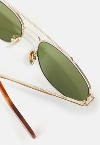 Ochelari de soare din metal auriu cu lentile verzi. Profil subțire cu rame rotunde, având un braț cu brand și un accent maro cu aspect de țestoasă.