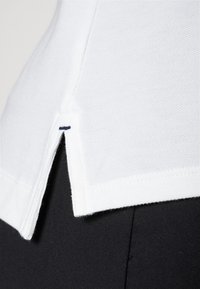 Bílá bavlněná košile s texturovaným povrchem, vybavená bočním rozparkem a modrým akcentním stehem na lemu, doplněná černou látkou.