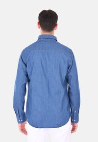 Camicia in denim blu, maniche lunghe, colletto classico, taglio dritto con polsini abbottonati. La vista posteriore mostra una trama liscia e dettagli di cucitura.