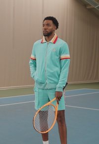Türkiissinine treeningülikond punaste ja kollaste triipudega, lühikeste varrukatega ning tõmblukuga ees. Hoidmas oranži tennisereketit väljakul.