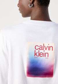 Felpa bianca con un grande motivo sulla schiena e colori in gradiente che vanno dal rosa al blu, e il testo "calvin klein" in rosso.