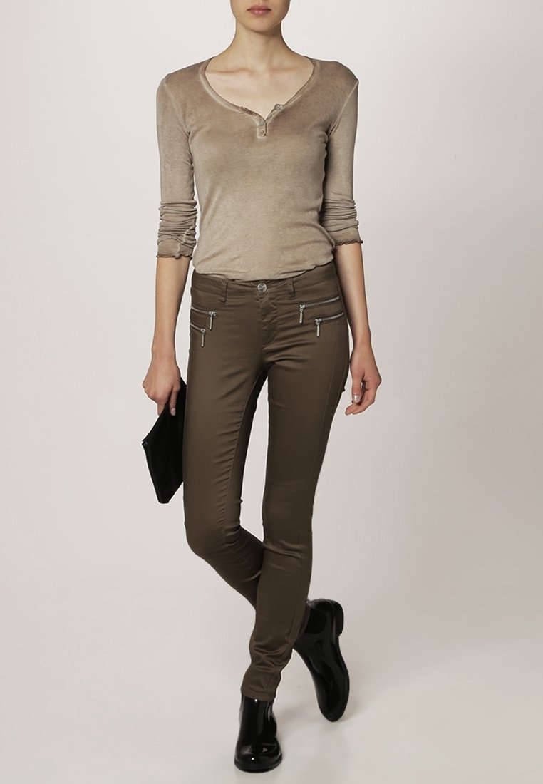 Femme portant un chemisier henley à manches longues beige, un pantalon skinny marron avec poches zippées, des bottines noires, tenant une pochette noire.