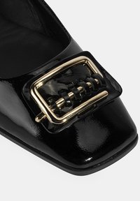 Glänzender schwarzer spitzer Schuh mit einer großen dekorativen goldenen und schwarzen quadratischen Schnalle auf der Vorderseite.