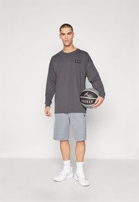 Grå langermet skjorte med en teksturert etikett, sammen med lysegrå shorts. Modellen holder en basketball, iført hvite treningssko og sokker.