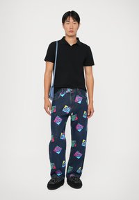Blugi din denim închis cu motive grafice vibrante în nuanțe de roz, albastru și verde. Cămașă polo neagră, geantă crossbody albastră și încălțăminte neagră.