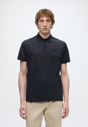 Ung mand med krøllet hår iført en sort poloshirt med korte ærmer og beige bukser, står foran en simpel lys baggrund.