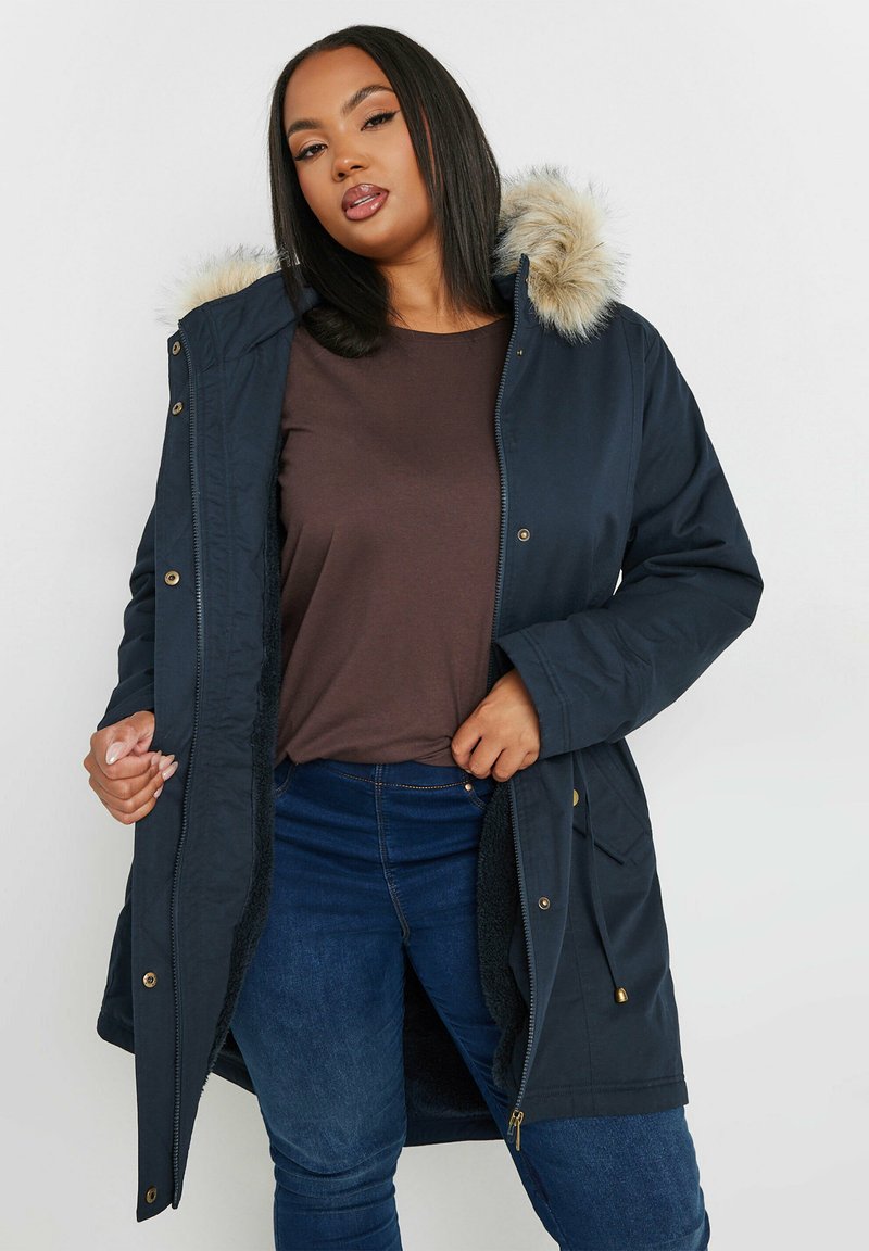 Marinfärgad parka-jacka med pälsförsedd huva, dragkedja och tryckknappar, fickor och mjukt fleecefoder. Bärs över en brun topp och blå jeans.