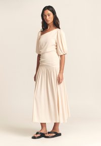 Lichtbeige jurk met pofmouwen, asymmetrische halslijn en geplooide taille. Rok is vloeiend en kniehoog. Gecombineerd met zwarte sandalen.