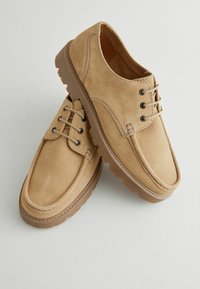 Zapatos de ante beige con un perfil bajo, puntera redondeada y diseño de cordones. Suela de goma estriada con acentos de doble costura.